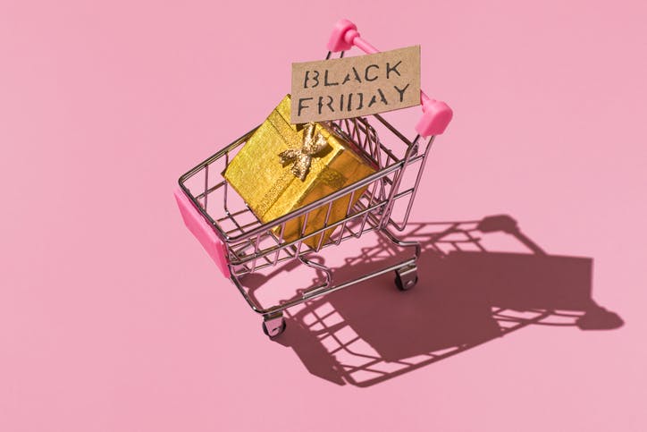 Metal shopping cart with black friday sign and golden gift box on pink background.