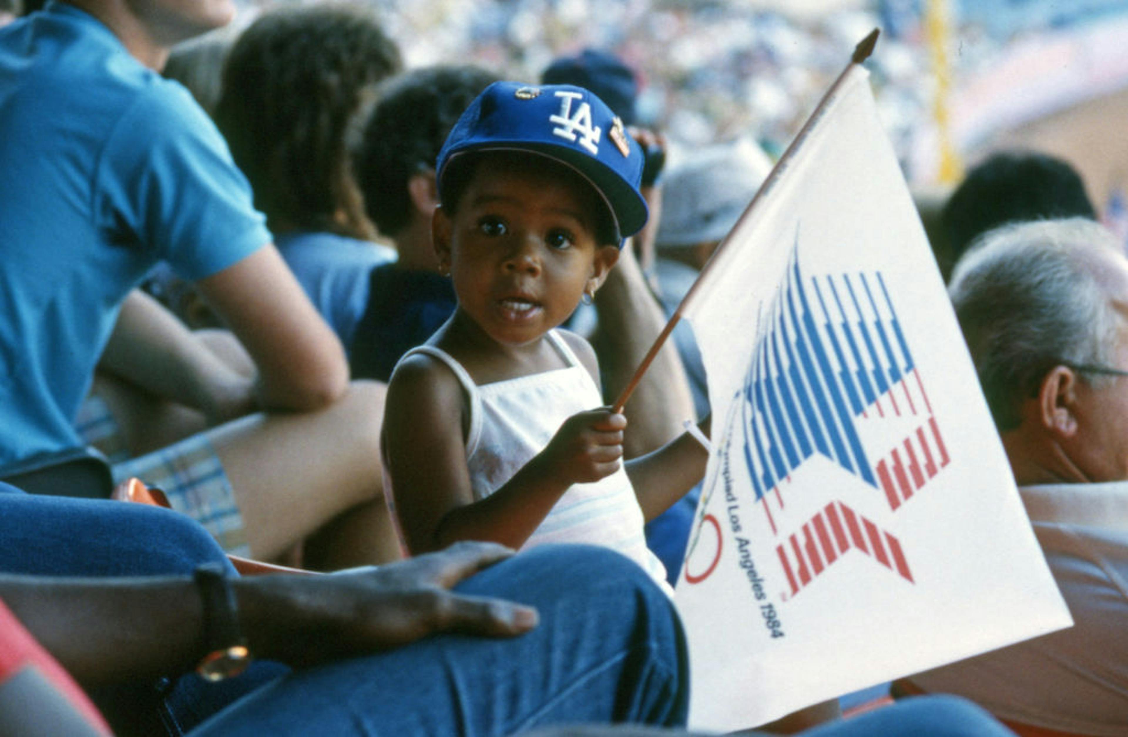 A small child wearing a Dodgers baseball cap and holding a 1984 Summer Olympics flag