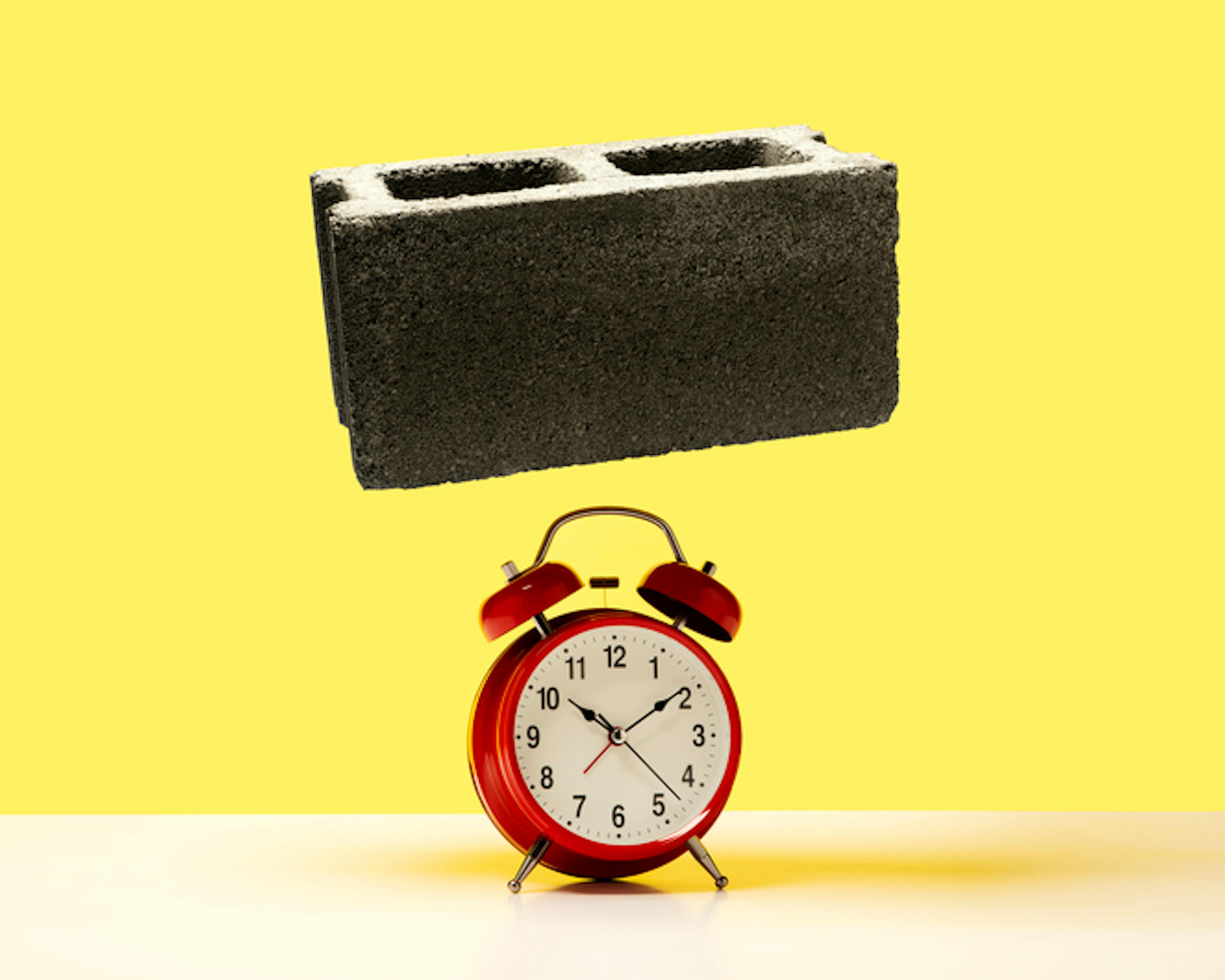 Cinder block about to fall on red alarm clock on white surface, yellow background