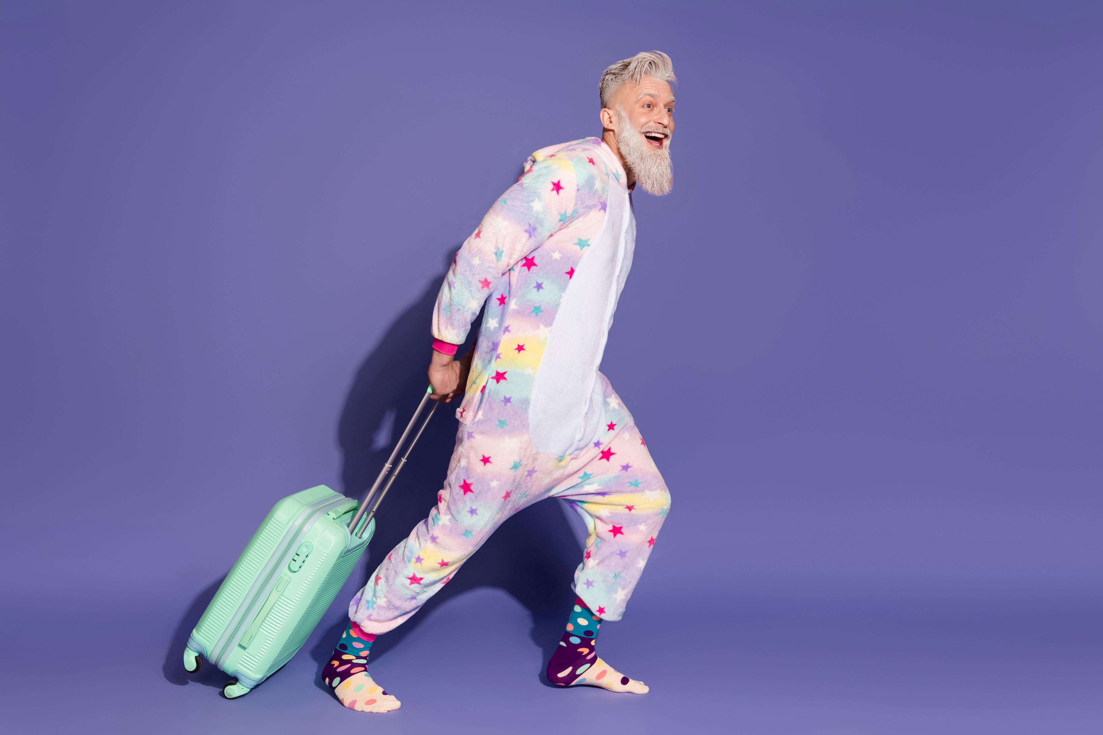 Joyful man in colorful unicorn pajamas with a suitcase against a violet background