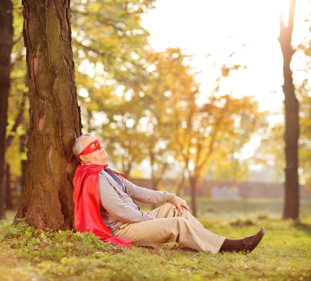 Senior in superhero outfit leaning on tree in park